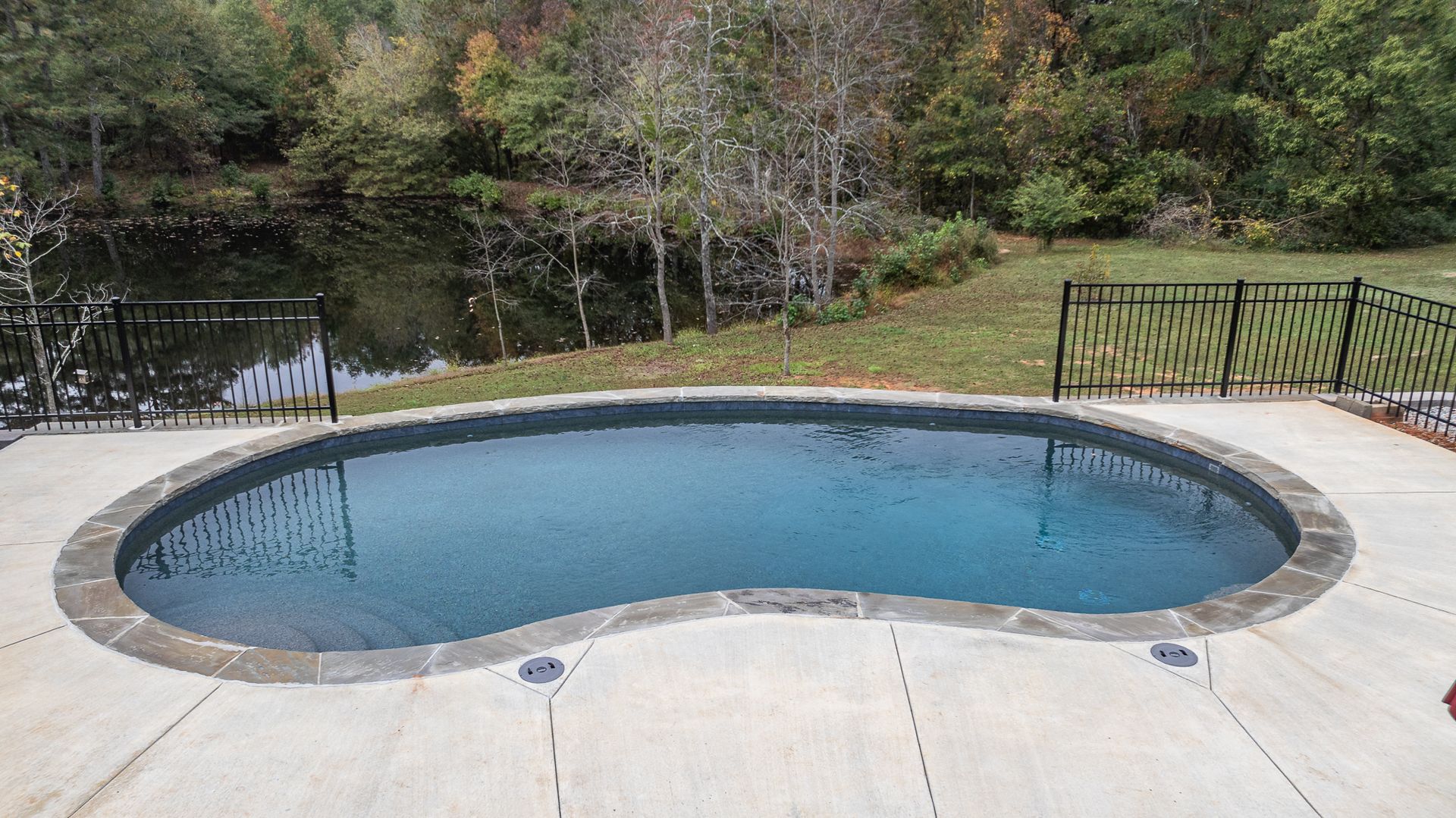 A large swimming pool is surrounded by trees and a fence.