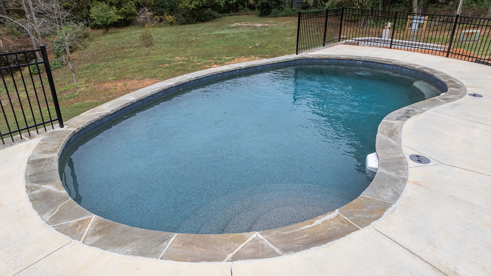 A large oval swimming pool is surrounded by a fence and a concrete patio.