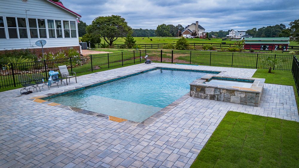 Rectangular pool with hot tub, patio, and fenced yard overlooking a field and houses on a cloudy day.