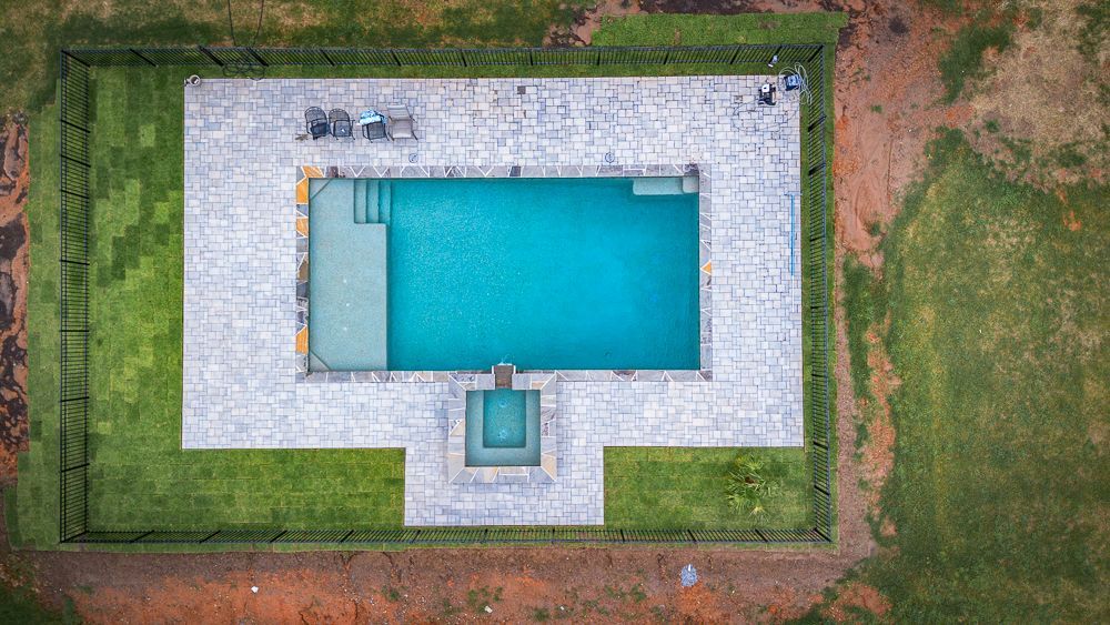 Aerial view of a rectangular pool with teal water, stone surround, and a small hot tub, fenced in with grass perimeter.