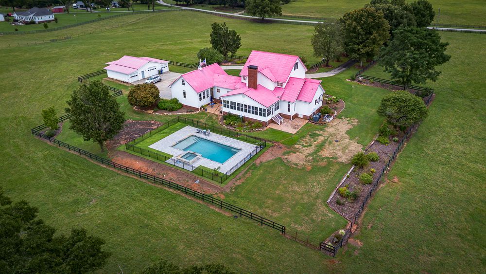 Aerial view of a white house with a pink roof, pool, and outbuildings surrounded by green grass.
