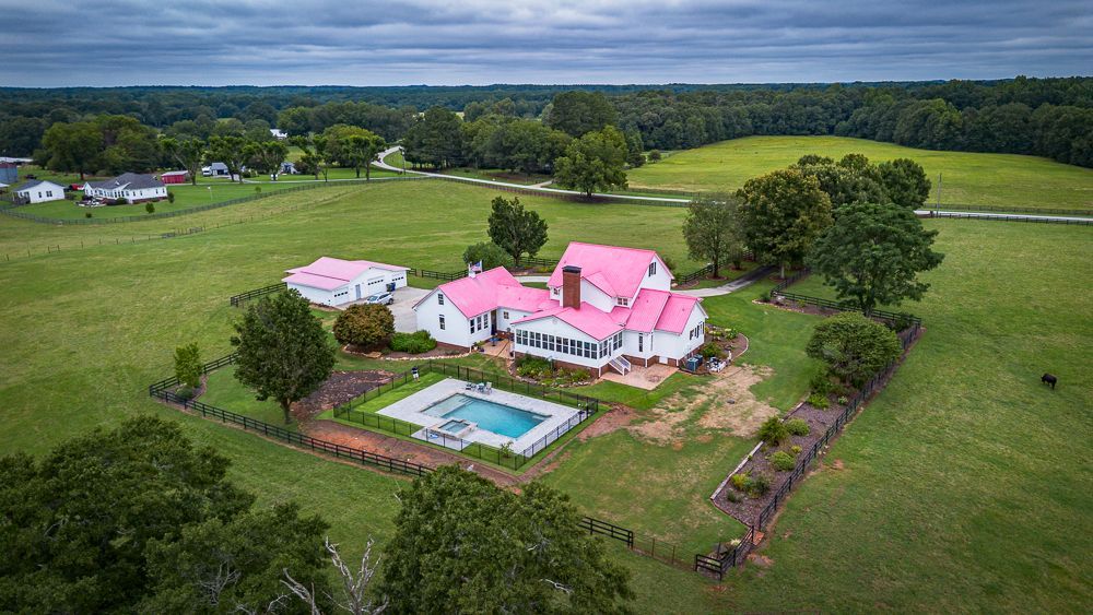 White farmhouse with pink roof, pool, and large green lawn, surrounded by trees and fields.