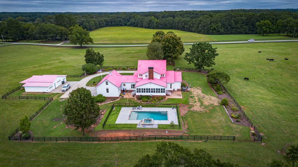 White farmhouse with pink roof and outbuildings, surrounded by green fields and a pool.