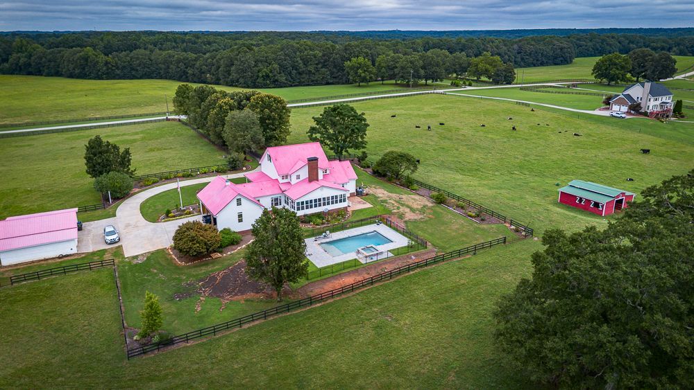 Aerial view of a white farmhouse with a pink roof, pool, and red barn on a green pasture.