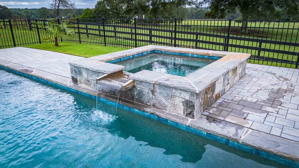 A backyard pool with a connected square hot tub, with water cascading from the hot tub.