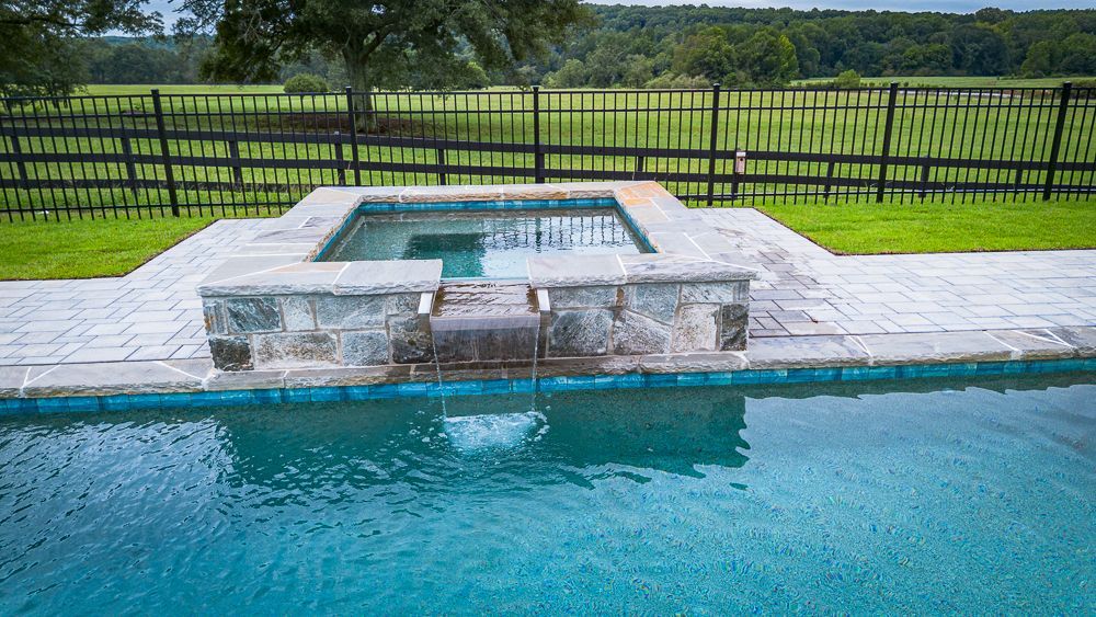 Swimming pool with spa, waterfall feature, and black fence in a grassy field.