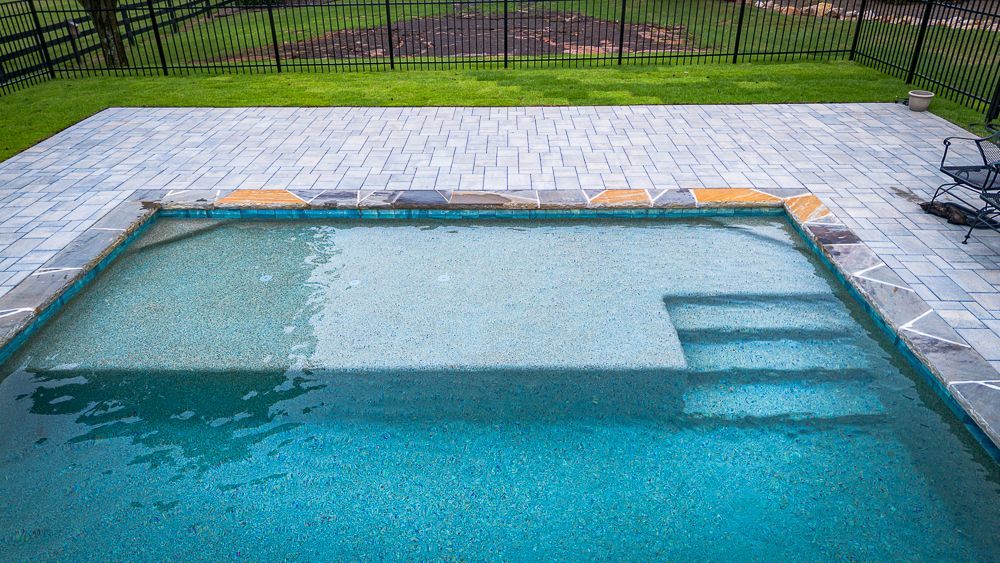 Blue pool with steps, surrounded by gray pavers and green grass, black fence in background.