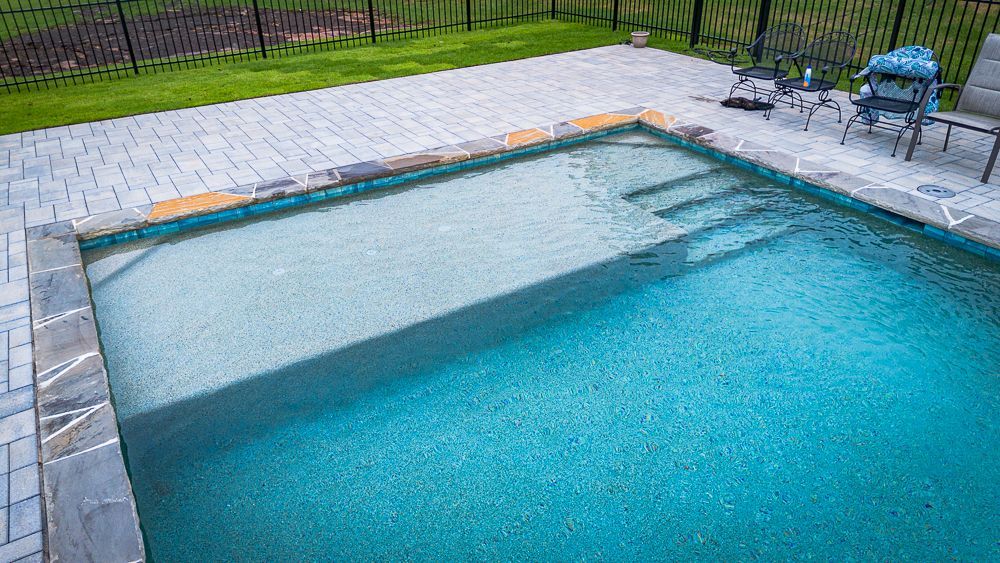 A rectangular swimming pool with blue water. Gray brick patio surrounds it, along with green grass.