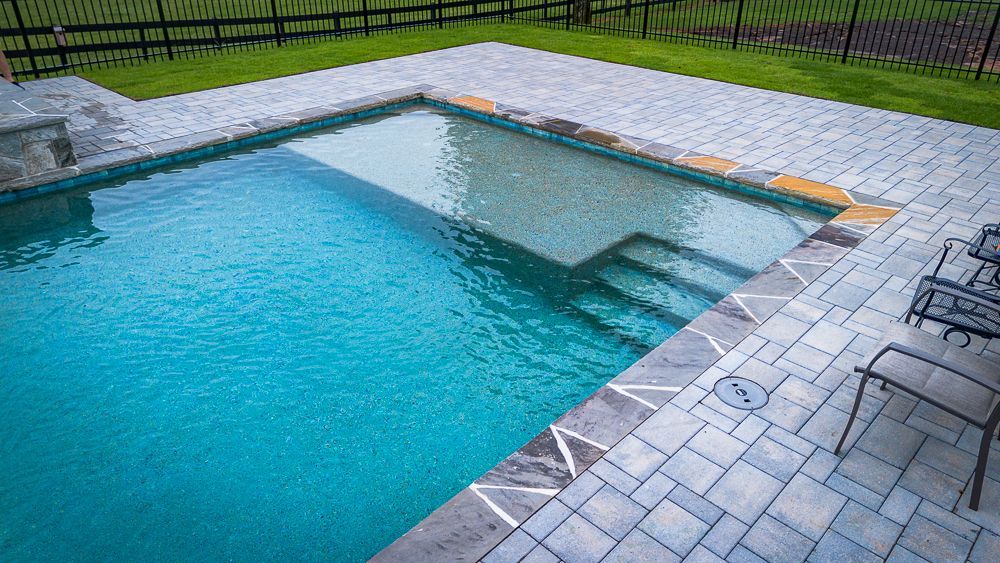 A rectangular swimming pool with gray brick patio, grass, and a fence in the background.