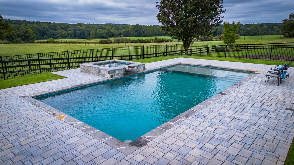 Rectangular pool with turquoise water, a small spa, surrounded by brick pavers, and a field in the background.