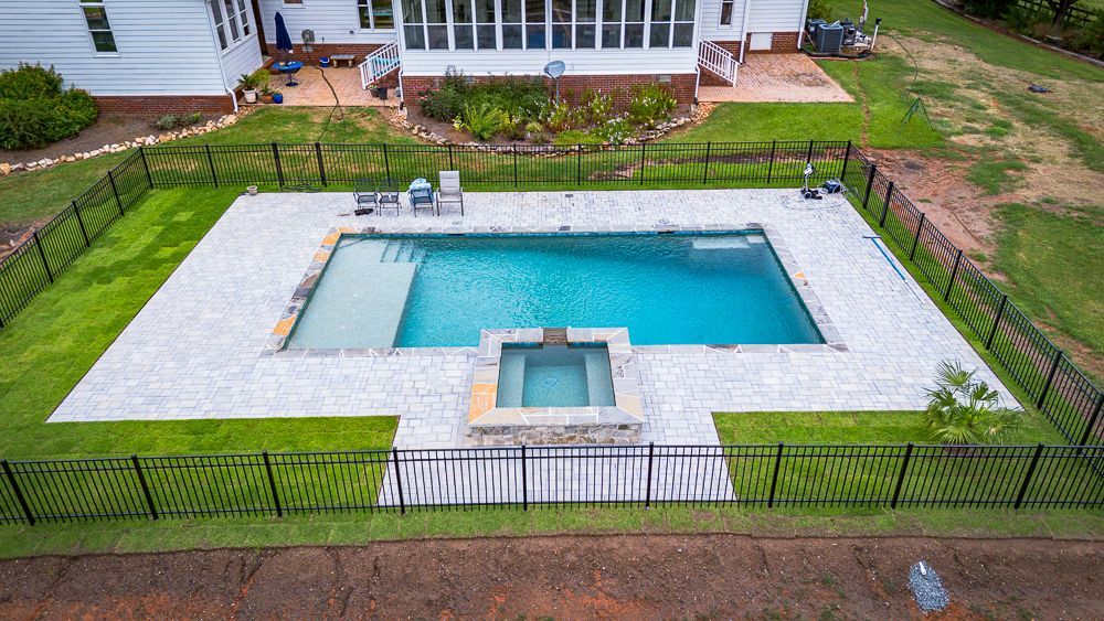 Rectangular pool with jacuzzi, surrounded by light stone, grass, and black fence in backyard.