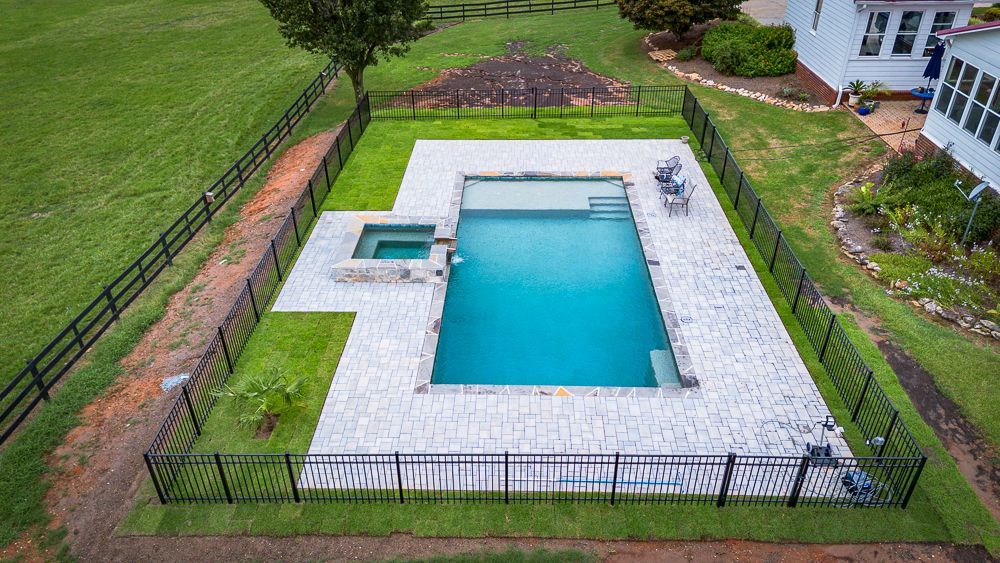 Aerial view of a rectangular pool and spa, surrounded by stone pavers and black fencing on green grass.
