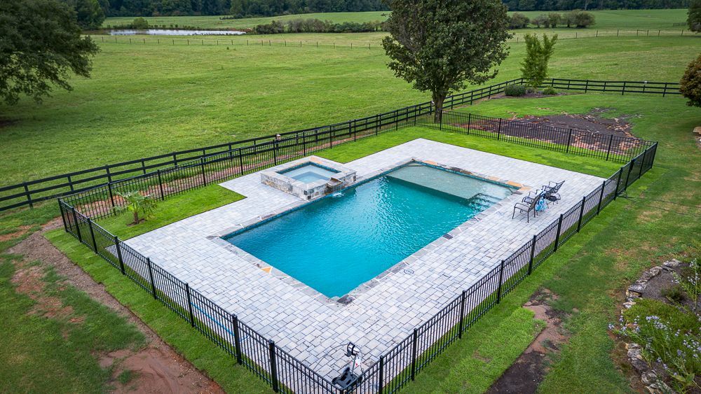Aerial view of a rectangular swimming pool with a spa, surrounded by grass and a black fence in a green field.