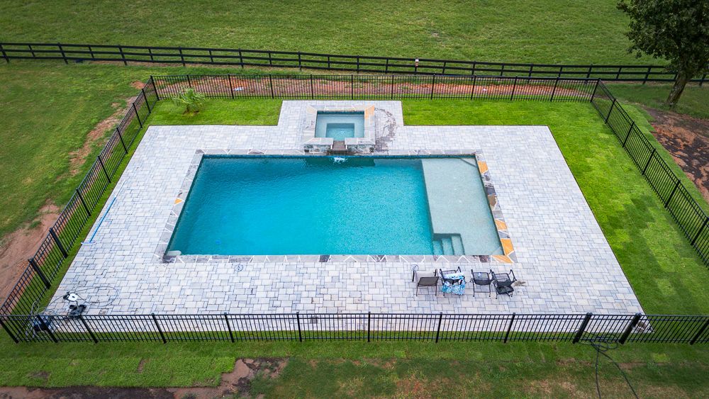 Aerial view of a rectangular pool with a connected hot tub surrounded by light stone and a black fence in a grassy yard.