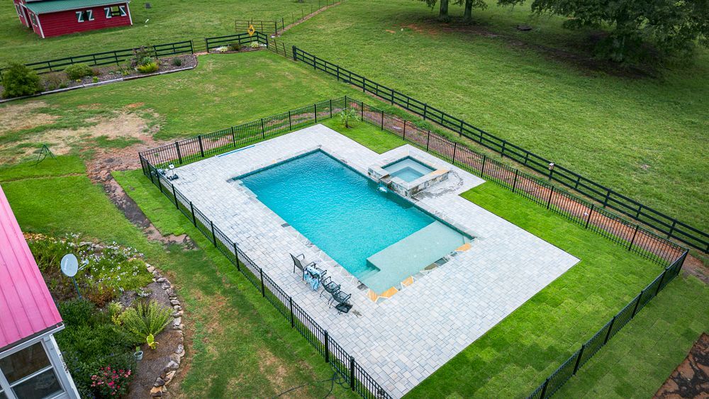Aerial view of a rectangular swimming pool with a connected spa. Surrounded by a brick patio and black fence on a green lawn.