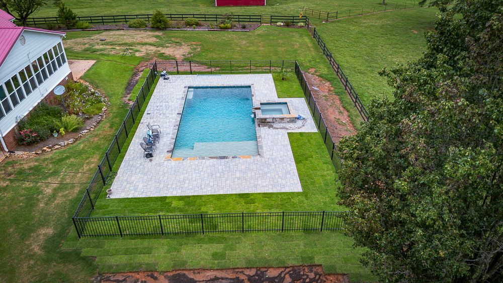 Aerial view of a pool and spa area surrounded by a fence and green grass, next to a house.