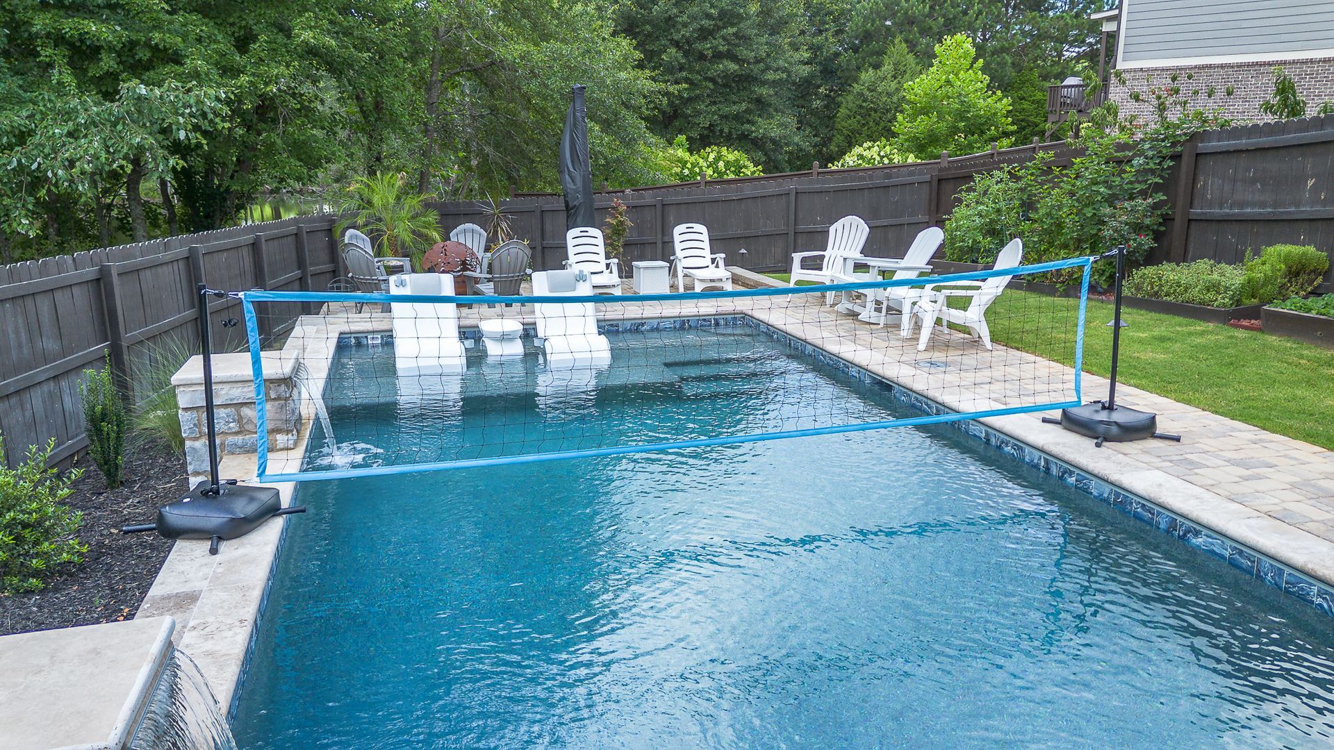 Blue pool with a volleyball net set up; white chairs surround the pool on a stone patio.