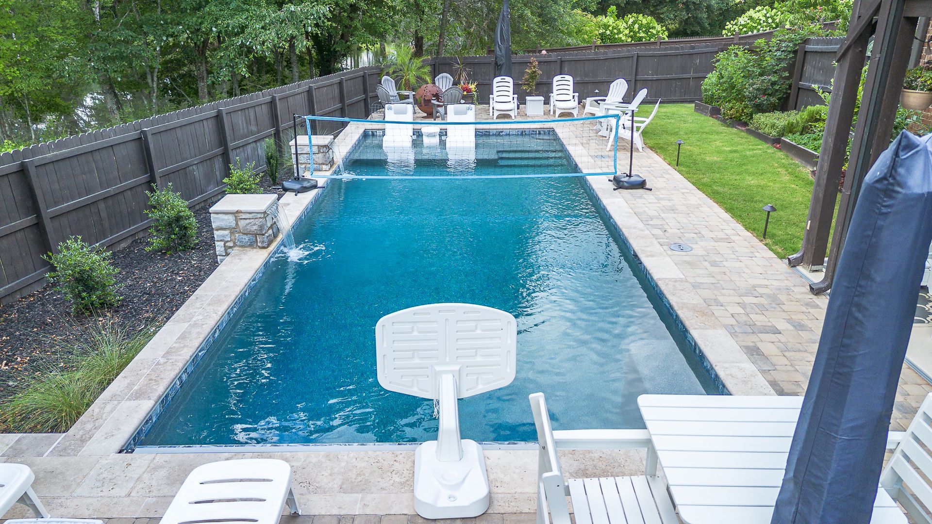 Rectangular outdoor pool with blue water, white chairs, and a basketball hoop, surrounded by a patio, grass, and a wooden fence.