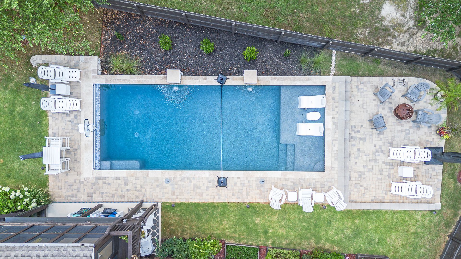 Overhead view of a rectangular pool with lounge chairs, surrounded by a patio, lawn, and a fire pit.