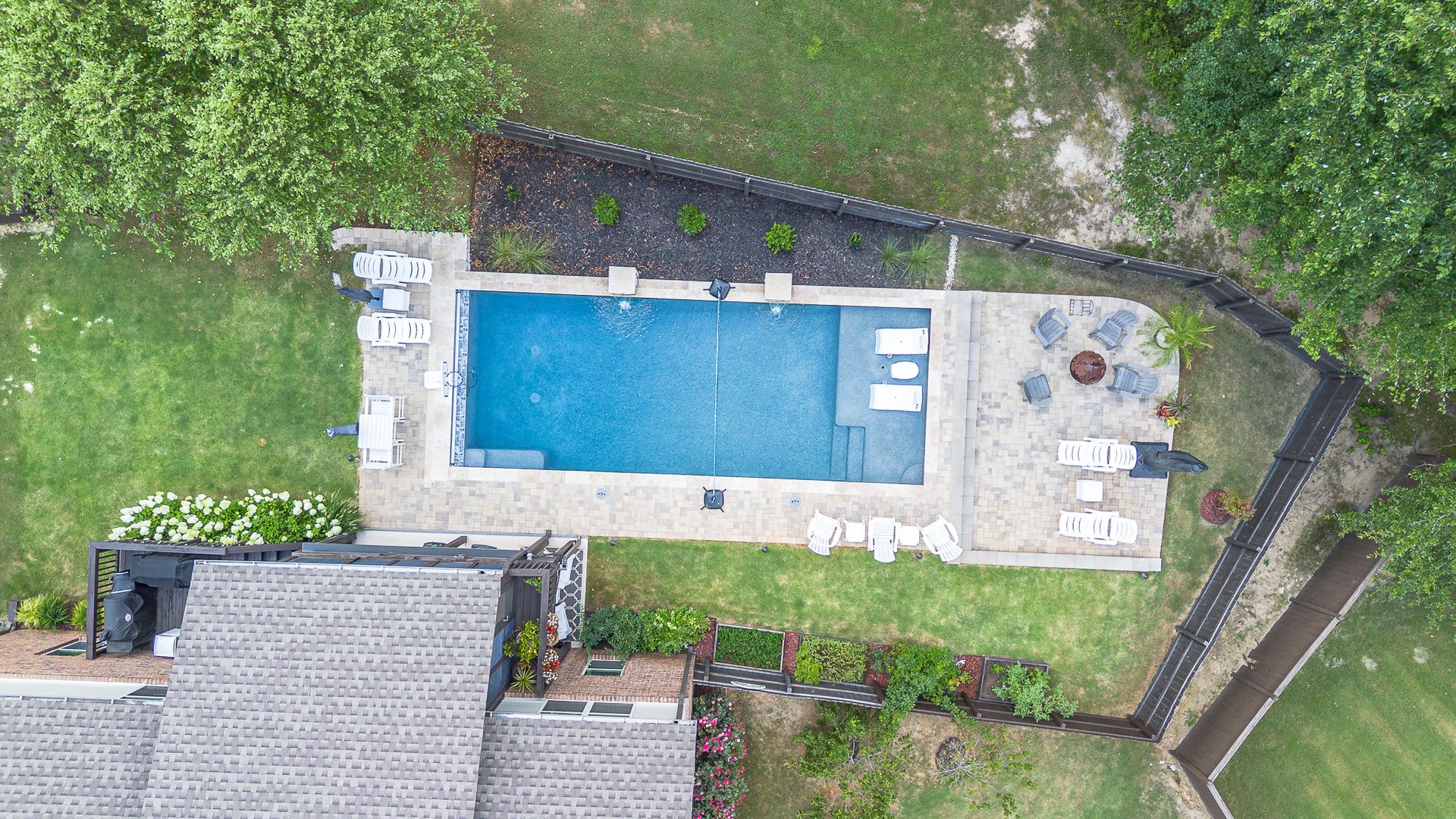 Aerial view of a pool surrounded by grass, with lounge chairs, a house, and trees.