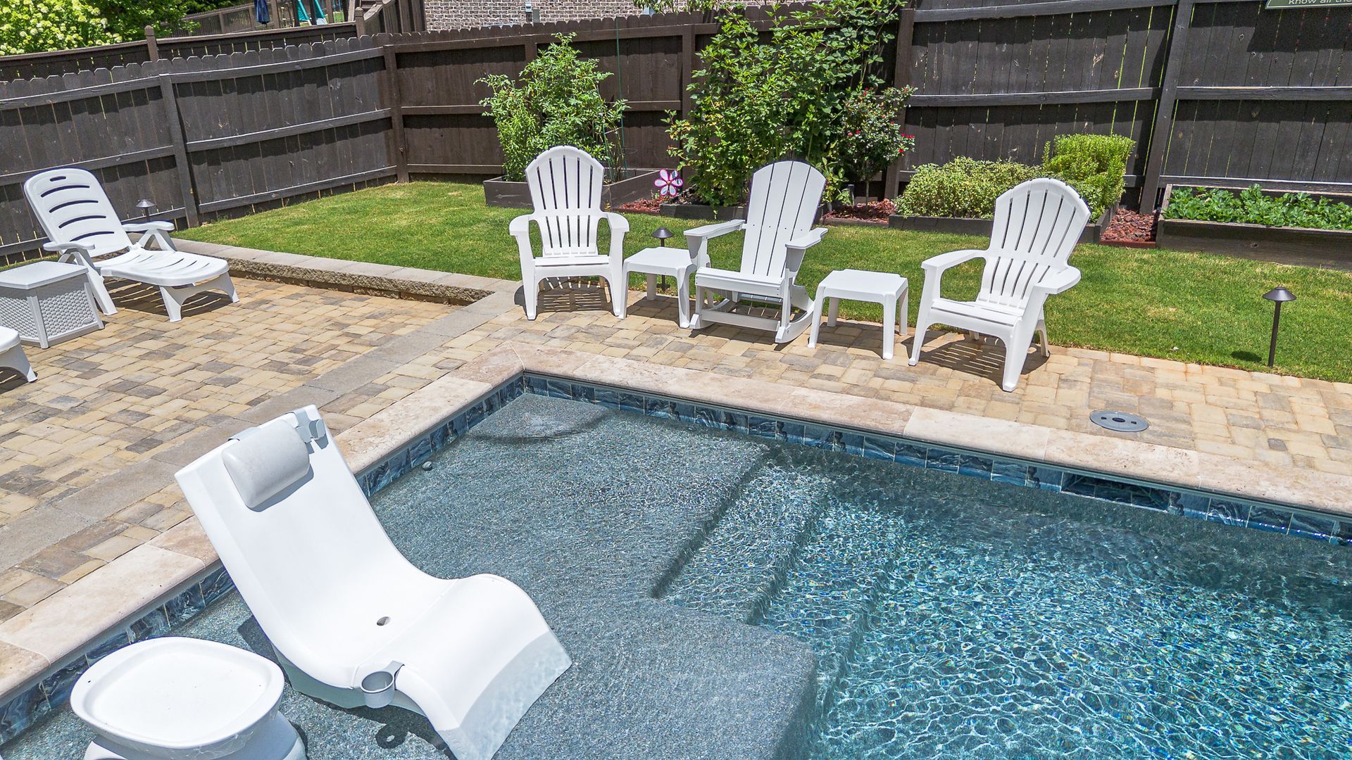 Poolside scene: white chairs and a lounge chair surround a pool with blue tile. Green grass, brown fence.