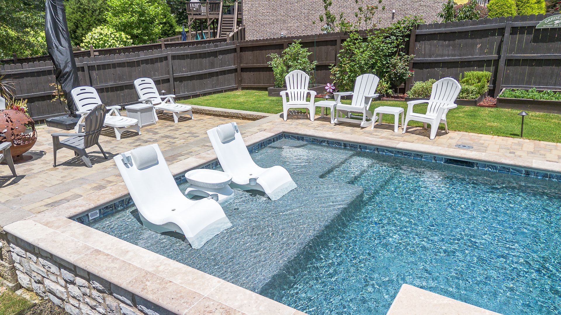 Backyard pool with white lounge chairs in the water and on the patio, surrounded by a fence and greenery.
