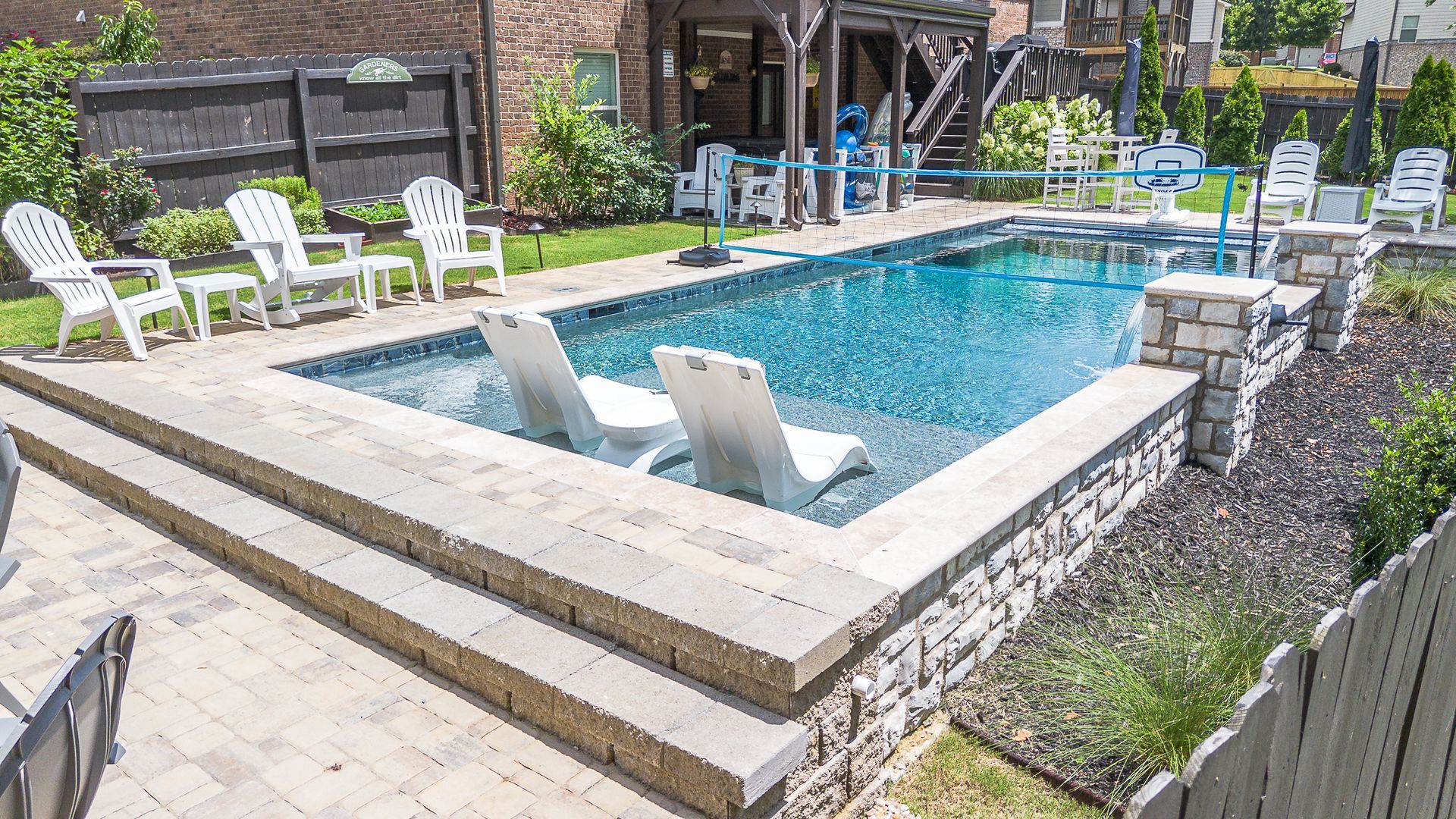 Rectangular swimming pool with white lounge chairs. Stone steps lead to a patio with white chairs; a wooden structure in the background.