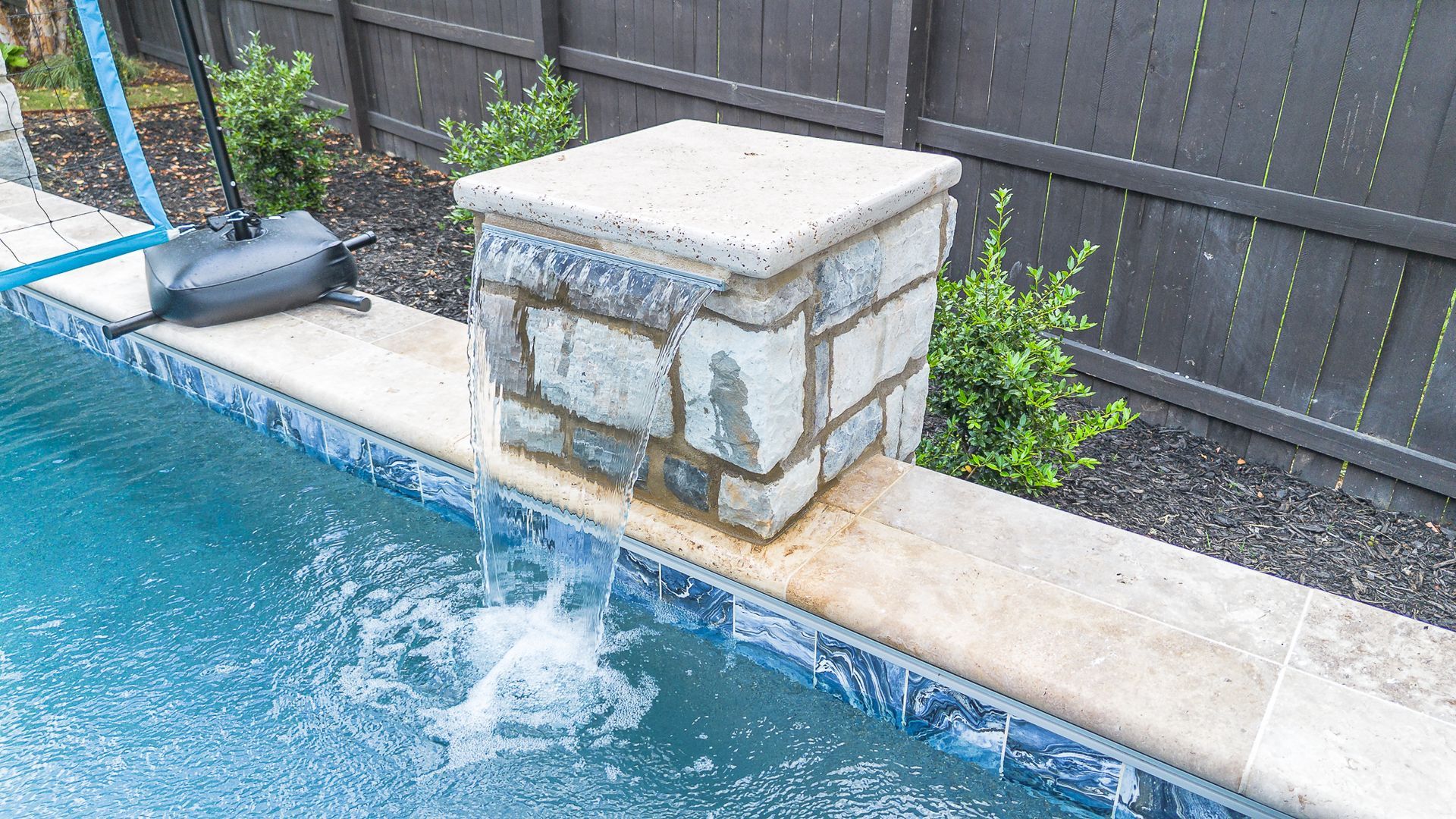 A pool with stone fountain, blue tile, and a dark wooden fence.
