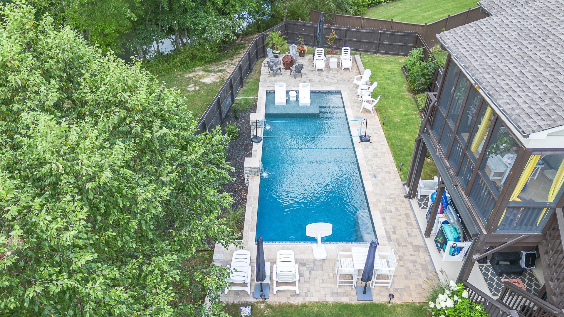 Aerial view of a rectangular pool with white chairs, next to a house with a screened porch and lush greenery.