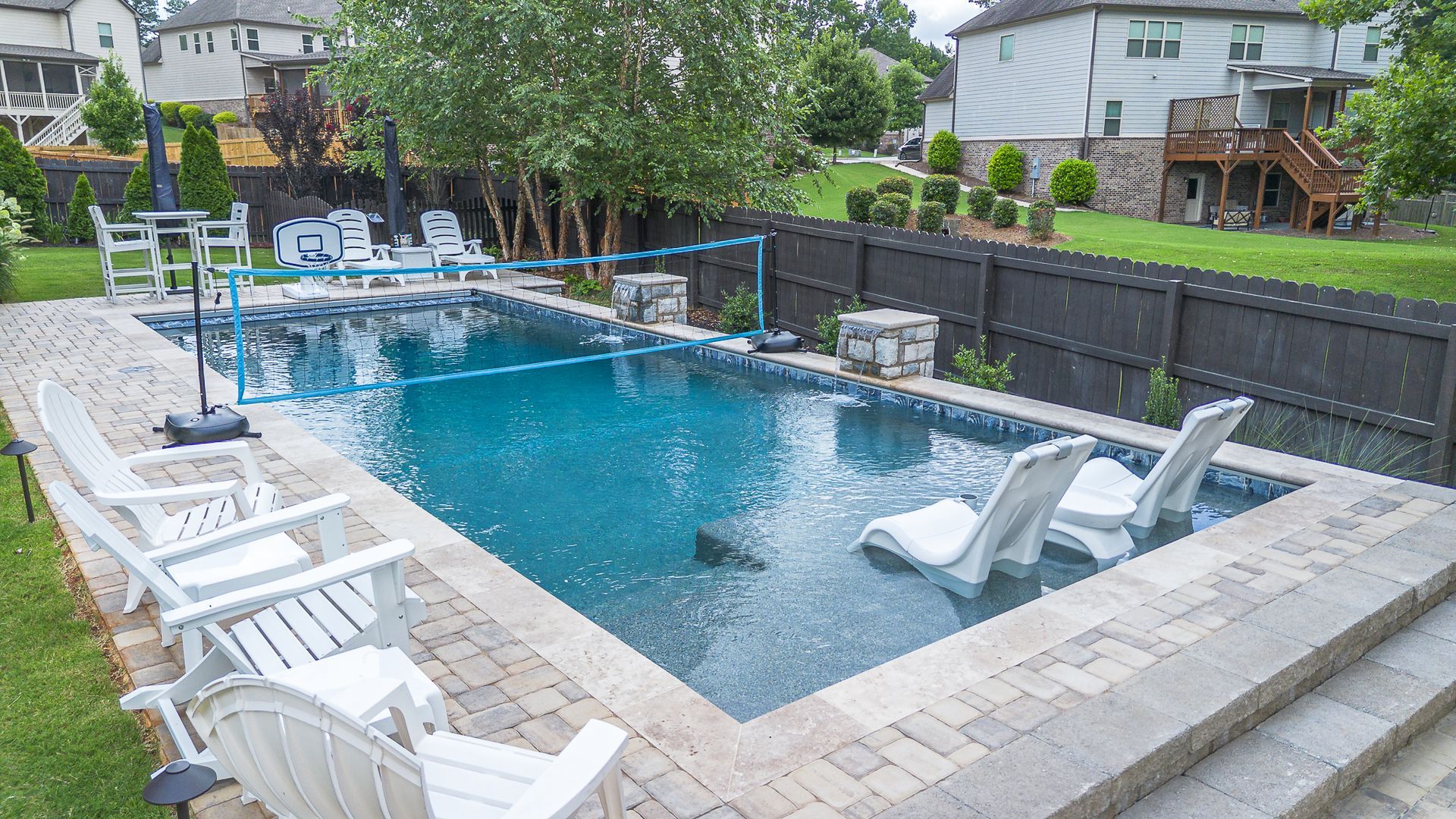Backyard pool with white chairs, surrounded by pavers and grass, with houses in background.