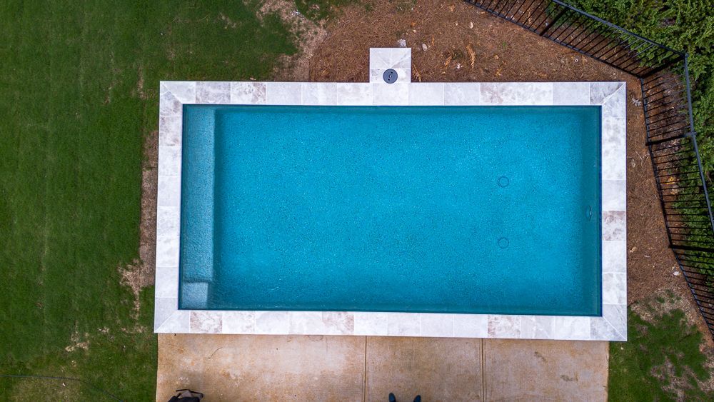 Overhead view of a rectangular turquoise pool with a light-colored stone border surrounded by grass and a fence.