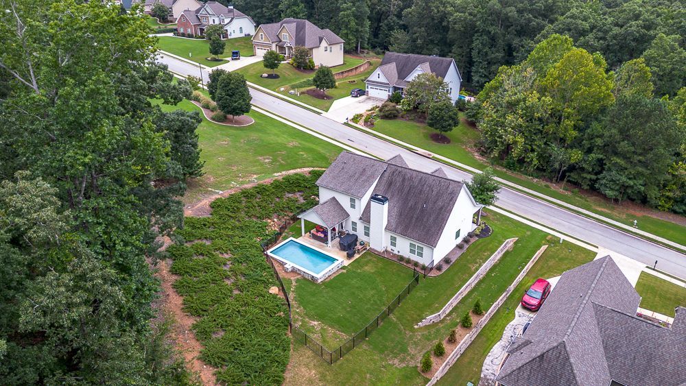 Aerial view of a white house with a pool, next to a road and other houses, surrounded by trees.