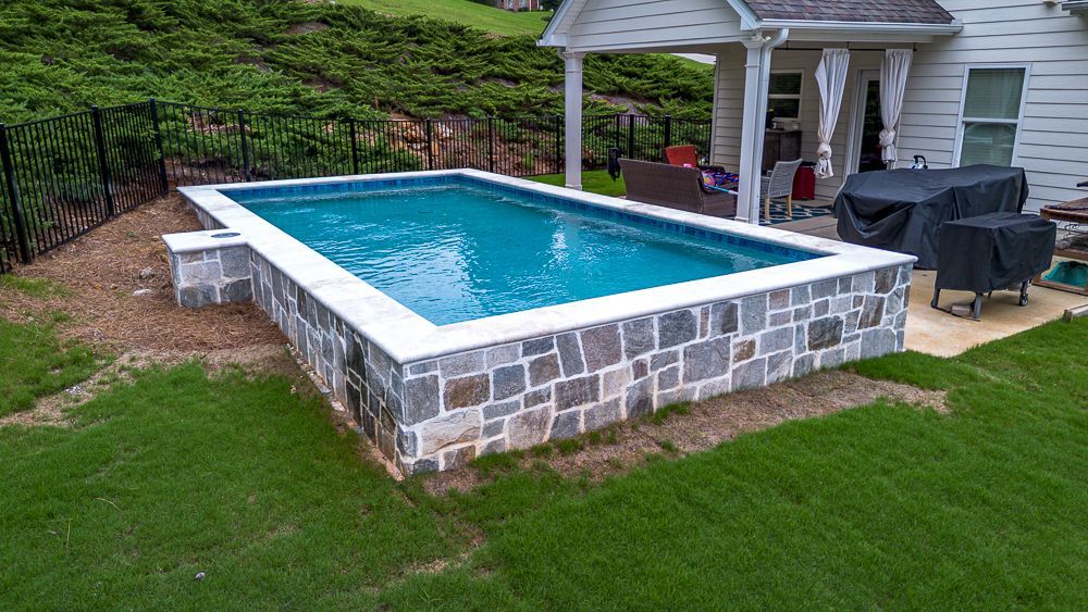 Rectangular in-ground pool with stone border, adjacent to a house patio. Green grass and landscaping surround it.