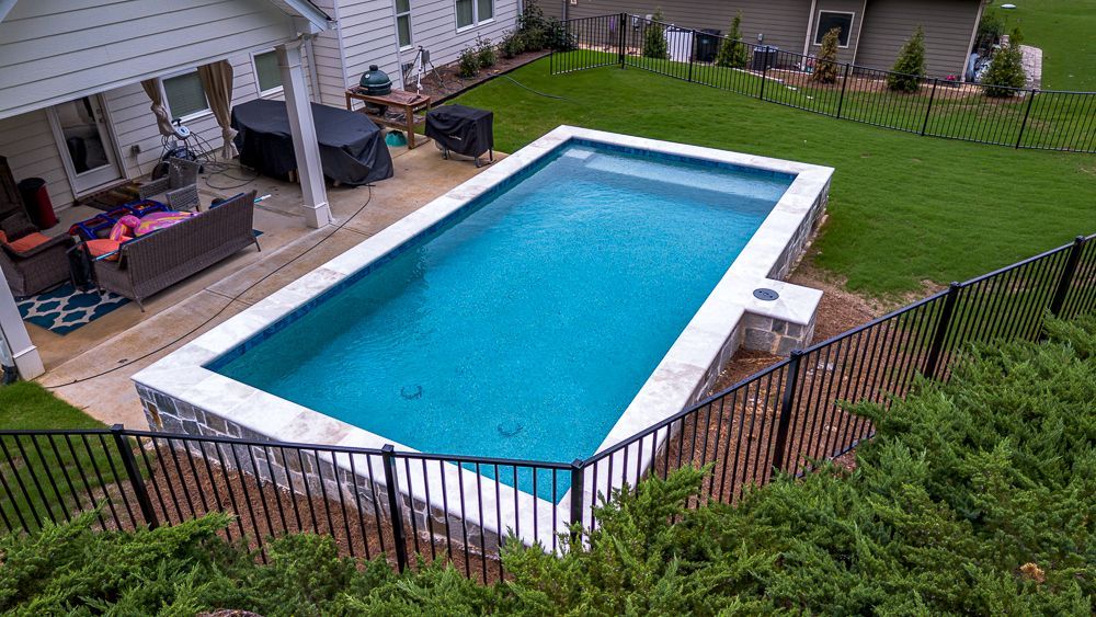 Rectangular swimming pool with blue water, next to a house, surrounded by a stone border and a black fence.