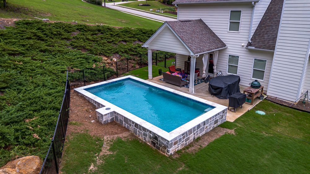 A rectangular pool with a white border in a backyard next to a house with a patio.