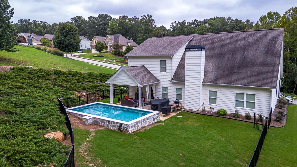 Backyard of a white house with a pool, green lawn, and trees on a hillside.