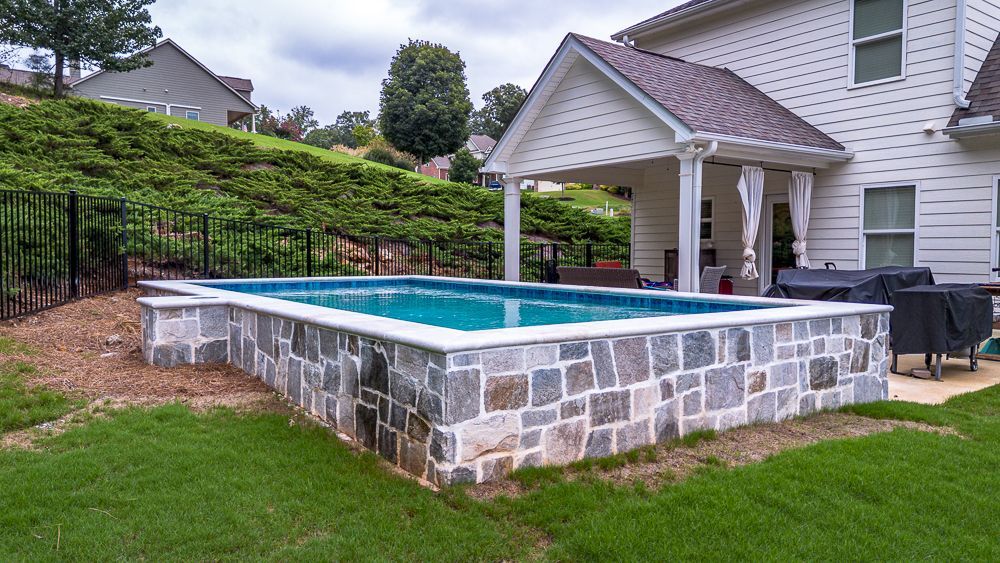 A rectangular pool with stone walls, surrounded by grass, next to a house with a covered patio.