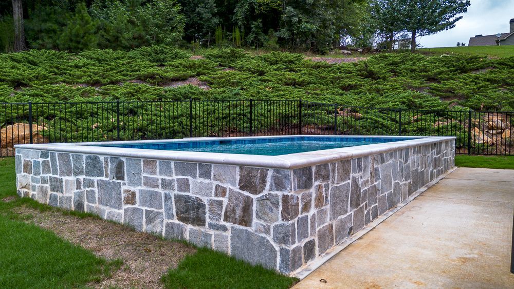 Rectangular stone-walled pool with blue water, set on a concrete patio with a grassy lawn. Trees in the background.