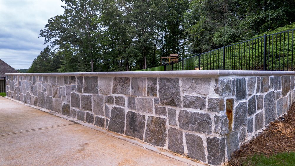 Stone retaining wall with a concrete cap. Brown and grey stone, green trees, and black fence in the background.
