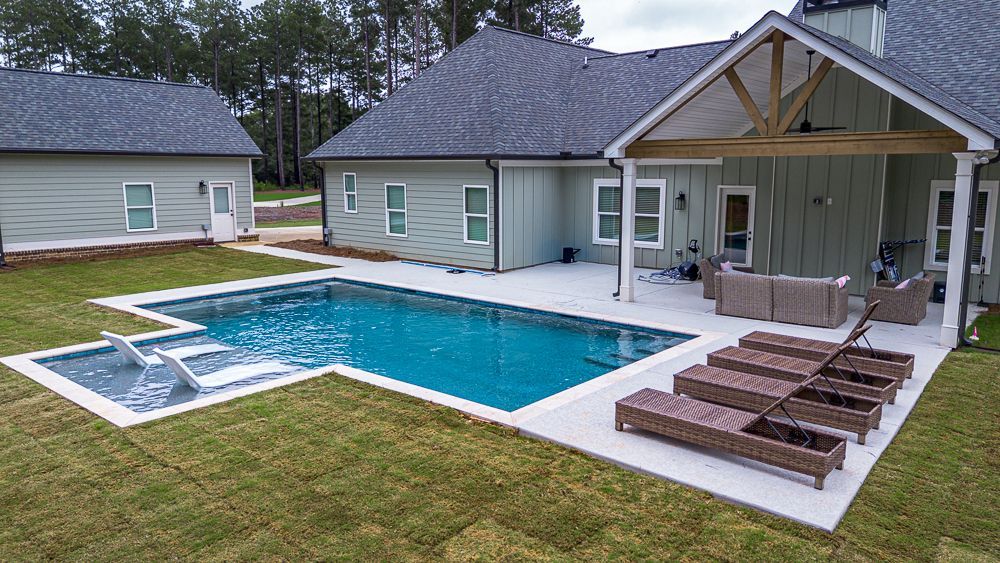 A modern house with a pool and patio in the backyard. Teal water, brown lounge chairs, and green grass.