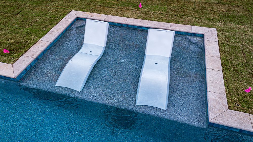 Two white lounge chairs submerged in a pool, next to a grassy lawn, with a blue water background.