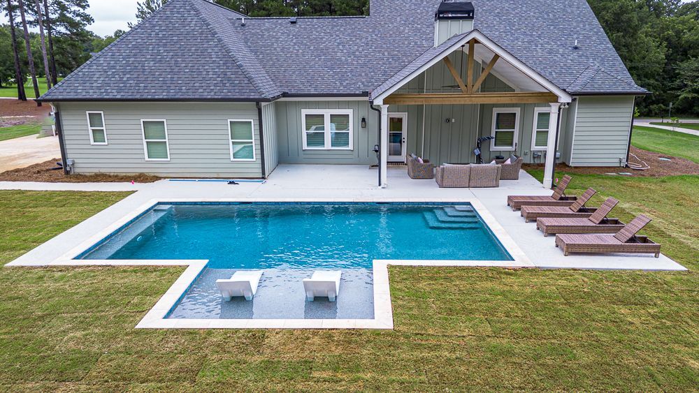 Backyard with a pool, patio, and house. The pool is blue, surrounded by gray concrete. There are lounge chairs.