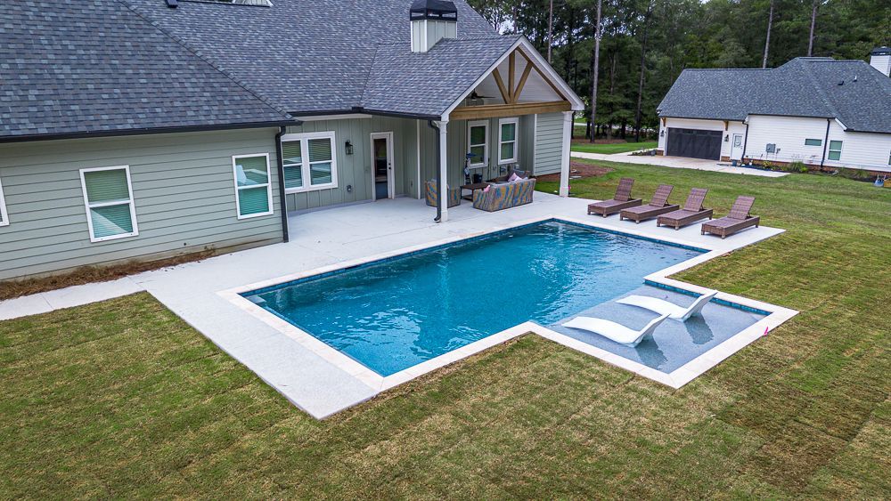 Backyard with a blue rectangular pool, tanning chairs, and a house with a covered patio.