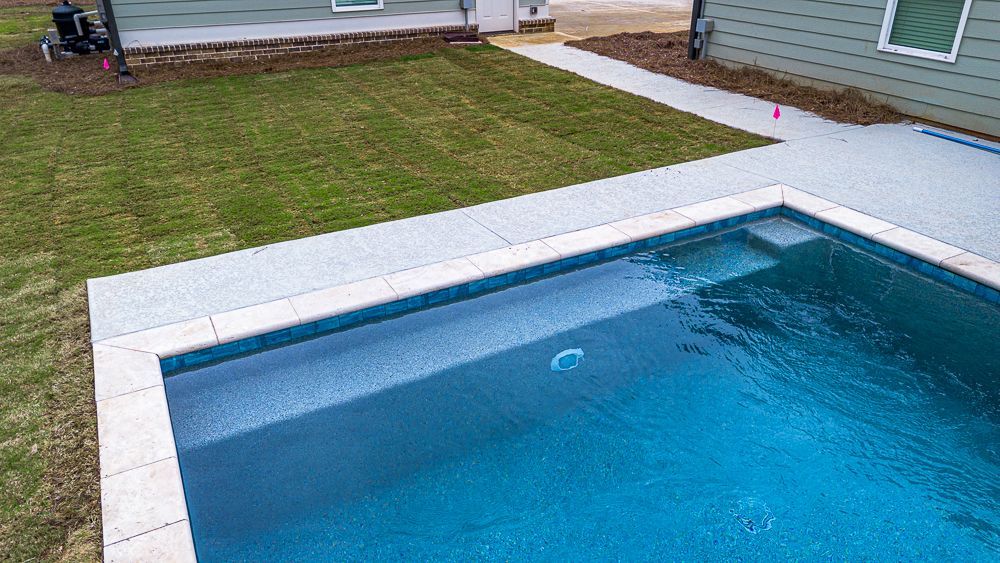 A rectangular swimming pool with blue water, surrounded by a concrete patio and green grass.