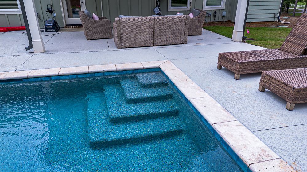 A backyard pool with steps, surrounded by a light concrete patio and wicker furniture.