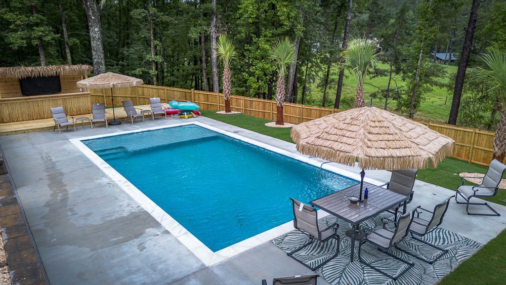 Backyard pool with a patio, umbrellas, and seating; surrounded by trees and a wooden fence.