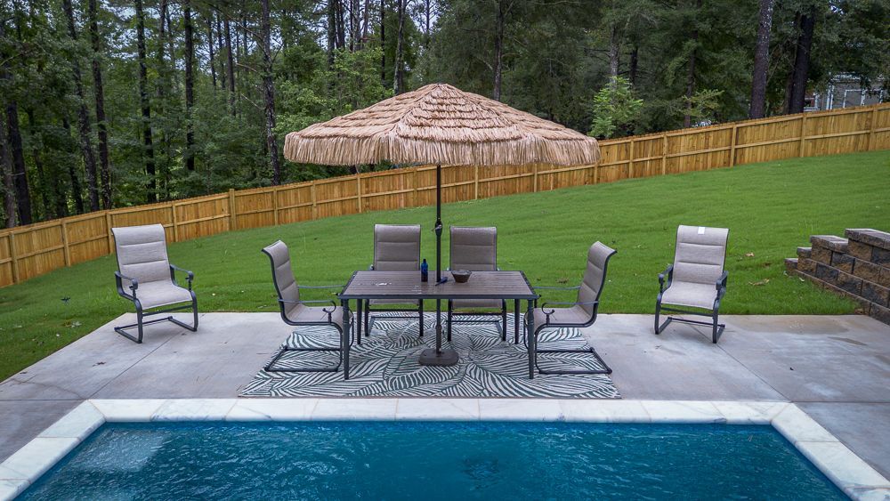 Backyard patio with pool, table, chairs, and umbrella; overlooking a fenced yard.