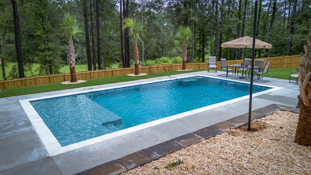 Backyard pool with patio furniture, surrounded by lawn, trees, and a wooden fence.