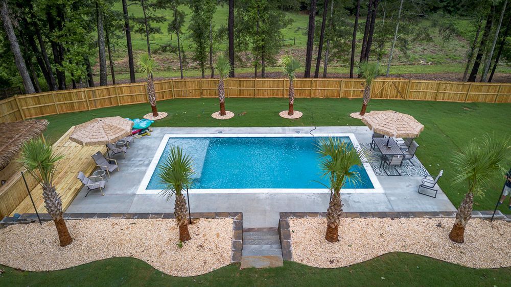 Pool area with palm trees, umbrellas, and a wooden fence, surrounded by grass and trees.
