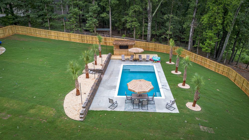Backyard with rectangular pool, tiki bar, and seating surrounded by a wooden fence and green grass.